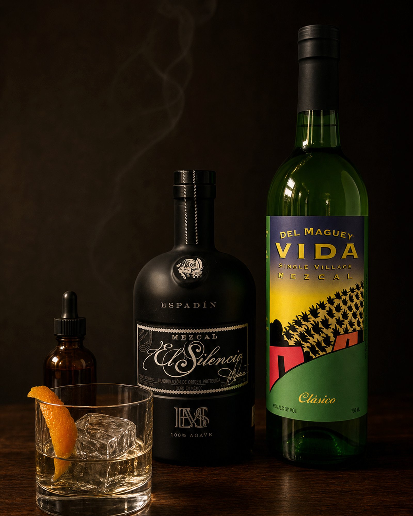 El Silencio and Del Maguey Vida Clásico mezcal bottles with a rocks glass, ice, orange twist, and bitters on dark walnut
