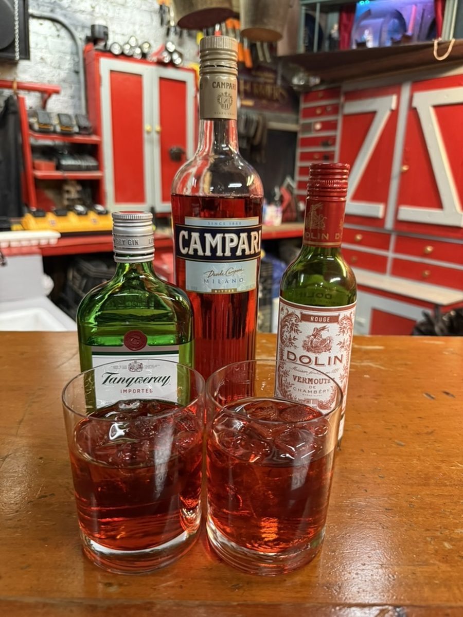 Two Negronis on a wooden bar at the Amateur Comedy Club, with bottles of Tanqueray gin, Campari, and Dolin Rouge vermouth behind them